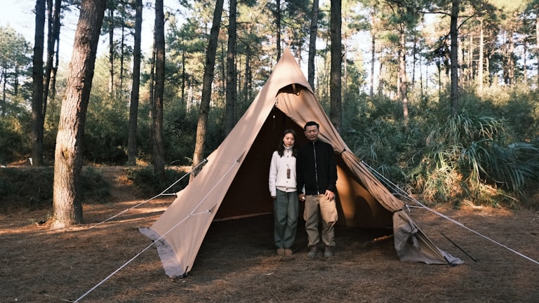 Two-person camping tent pitched in a lush forest clearing at sunrise.