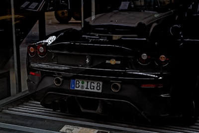 A black sports car is parked indoors on a metallic platform. The license plate indicates a European origin. The car's sleek design is highlighted by its low profile and round taillights. In the background, part of another classic car is visible, suggesting a garage or showroom setting.