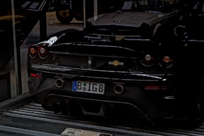 A black sports car is parked indoors on a metallic platform. The license plate indicates a European origin. The car's sleek design is highlighted by its low profile and round taillights. In the background, part of another classic car is visible, suggesting a garage or showroom setting.