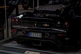 A black sports car is parked indoors on a metallic platform. The license plate indicates a European origin. The car's sleek design is highlighted by its low profile and round taillights. In the background, part of another classic car is visible, suggesting a garage or showroom setting.