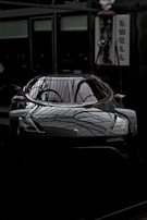 A sleek, modern car with a dark, reflective surface parked indoors. The lighting creates an intricate pattern on the car, highlighting its aerodynamic features. The setting appears to be a showroom or a garage with dim lighting.