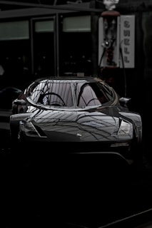 A sleek, modern car with a dark, reflective surface parked indoors. The lighting creates an intricate pattern on the car, highlighting its aerodynamic features. The setting appears to be a showroom or a garage with dim lighting.
