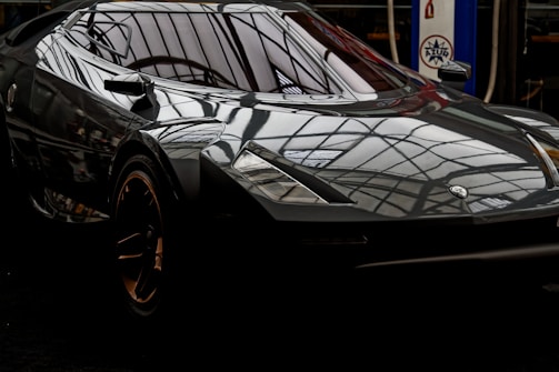 A sleek black sports car with a reflective surface showcasing a geometric pattern from the surrounding ceiling. The car has a modern and angular design with gold accents on the rims. There is a logo on the front of the car. In the background, a vintage-looking gas pump can be seen with a distinct logo and signage.