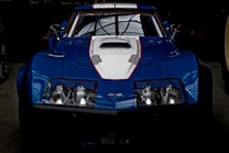 A sleek blue sports car with a white hood and red accent lines. The car features a distinctive design with a prominent front grille and round headlights. The vehicle has a low profile, emphasizing its aerodynamic shape, and is parked inside a dimly lit garage.