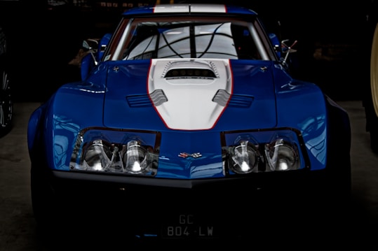 A sleek blue sports car with a white hood and red accent lines. The car features a distinctive design with a prominent front grille and round headlights. The vehicle has a low profile, emphasizing its aerodynamic shape, and is parked inside a dimly lit garage.