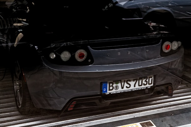 The rear view of a dark-colored sports car with the brand logo visible. The vehicle has European-style license plates and is parked indoors, possibly in a garage or showroom. The lighting casts shadows, creating a somewhat dramatic effect.