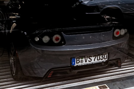 The rear view of a dark-colored sports car with the brand logo visible. The vehicle has European-style license plates and is parked indoors, possibly in a garage or showroom. The lighting casts shadows, creating a somewhat dramatic effect.
