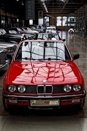 A bright red vintage convertible car is prominently positioned at the forefront of a collection of classic cars inside a spacious industrial-style showroom. The setting features large windows and high ceilings, creating an ambiance that highlights the car's sleek design and shiny exterior. Several other cars are aligned in neat rows behind the red car, showcasing a variety of classic and luxury models.