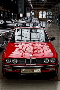 A bright red vintage convertible car is prominently positioned at the forefront of a collection of classic cars inside a spacious industrial-style showroom. The setting features large windows and high ceilings, creating an ambiance that highlights the car's sleek design and shiny exterior. Several other cars are aligned in neat rows behind the red car, showcasing a variety of classic and luxury models.