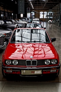 A bright red vintage convertible car is prominently positioned at the forefront of a collection of classic cars inside a spacious industrial-style showroom. The setting features large windows and high ceilings, creating an ambiance that highlights the car's sleek design and shiny exterior. Several other cars are aligned in neat rows behind the red car, showcasing a variety of classic and luxury models.