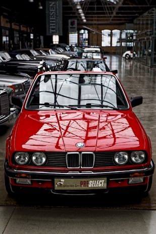 A bright red vintage convertible car is prominently positioned at the forefront of a collection of classic cars inside a spacious industrial-style showroom. The setting features large windows and high ceilings, creating an ambiance that highlights the car's sleek design and shiny exterior. Several other cars are aligned in neat rows behind the red car, showcasing a variety of classic and luxury models.