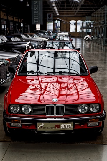 A bright red vintage convertible car is prominently positioned at the forefront of a collection of classic cars inside a spacious industrial-style showroom. The setting features large windows and high ceilings, creating an ambiance that highlights the car's sleek design and shiny exterior. Several other cars are aligned in neat rows behind the red car, showcasing a variety of classic and luxury models.