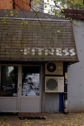 A building entrance with a door and windows is visible. Above the door, there is a sloped roof with the word 'FITNESS' written on it in white letters. An air conditioning unit is mounted next to the door. Leaves are scattered on the ground, and tree branches with leaves hang over the roof.