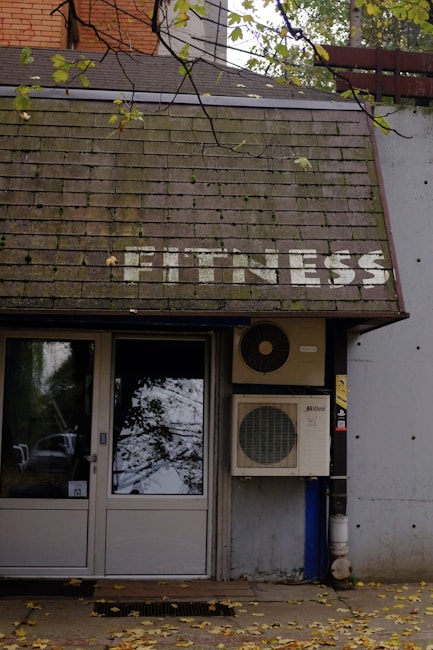 A building entrance with a door and windows is visible. Above the door, there is a sloped roof with the word 'FITNESS' written on it in white letters. An air conditioning unit is mounted next to the door. Leaves are scattered on the ground, and tree branches with leaves hang over the roof.