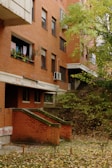 A brick apartment building with balconies, where laundry is hanging on one of the balconies, surrounded by green leafy trees. There are visible stairs leading to an entrance, and graffiti is seen on the lower part of the building.