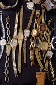 A collection of various vintage wristwatches with different designs and metal bands are laid out on a dark surface. The predominant colors are gold and silver, with intricate bracelet patterns and rectangular, oval, and round watch faces.