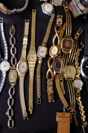 A collection of various vintage wristwatches with different designs and metal bands are laid out on a dark surface. The predominant colors are gold and silver, with intricate bracelet patterns and rectangular, oval, and round watch faces.