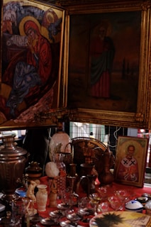 A collection of religious icons and vintage items is displayed, including framed paintings of religious figures with halos, various decorative vases, pitchers, and glassware on a red tablecloth. There is a central piece depicting a solemn religious figure with a halo, surrounded by intricate frames. The overall setup suggests a mix of spirituality and antique charm.
