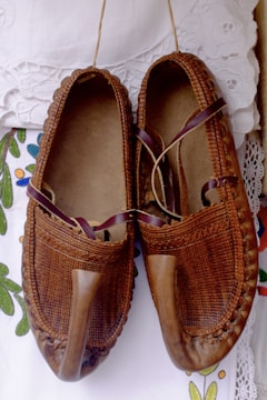A pair of traditional brown leather shoes with a pointed toe design, intricately woven details, and leather straps. They are placed on a white, lace-detailed fabric with colorful floral embroidery.