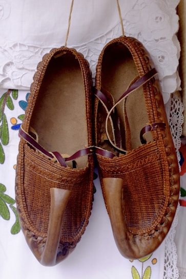 A pair of traditional brown leather shoes with a pointed toe design, intricately woven details, and leather straps. They are placed on a white, lace-detailed fabric with colorful floral embroidery.