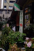An outdoor scene in front of a photo shop featuring a large Fujifilm sign. The storefront is surrounded by potted plants and flowers. There is also a Kodak sign on the building. The background includes an older building with visible electrical wires and laundry hanging to dry.