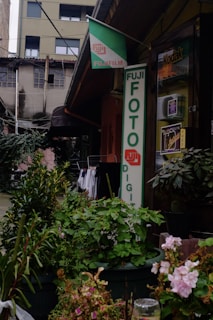 An outdoor scene in front of a photo shop featuring a large Fujifilm sign. The storefront is surrounded by potted plants and flowers. There is also a Kodak sign on the building. The background includes an older building with visible electrical wires and laundry hanging to dry.