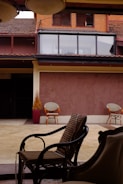 An outdoor patio area with three wicker chairs arranged under a covered section. The background includes a building façade with large windows and wooden shutters, featuring brown and reddish tones. A potted plant sits to the side, adding a touch of greenery.