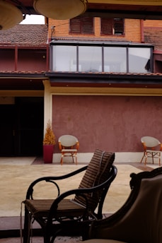 An outdoor patio area with three wicker chairs arranged under a covered section. The background includes a building façade with large windows and wooden shutters, featuring brown and reddish tones. A potted plant sits to the side, adding a touch of greenery.
