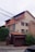 A multi-story residential house with a beige facade and reddish-brown accents. The house features numerous windows, some with shutters partially closed. There is a brick fence with a closed gate at the front. Several bushes and shrubs are visible around the house, and a series of power lines are prominently hanging across the upper part of the image.