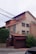 A multi-story residential house with a beige facade and reddish-brown accents. The house features numerous windows, some with shutters partially closed. There is a brick fence with a closed gate at the front. Several bushes and shrubs are visible around the house, and a series of power lines are prominently hanging across the upper part of the image.