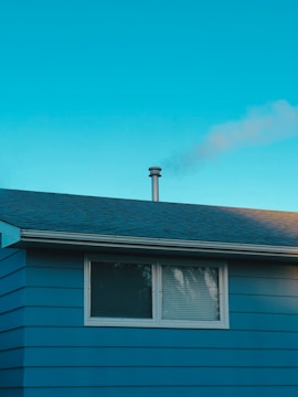 A residential house with blue siding and a sloped roof, along with a chimney emitting smoke. The window on the side of the house is closed with blinds. The sky is clear and blue, indicating bright weather.