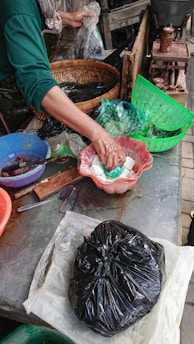 A person is preparing or handling food items at a market or stall. Various utensils and bowls are present, with the person using their hands to handle white pasty food in a red bowl. A black plastic bag and several baskets and containers are also visible in the scene.