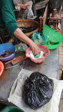 A person is preparing or handling food items at a market or stall. Various utensils and bowls are present, with the person using their hands to handle white pasty food in a red bowl. A black plastic bag and several baskets and containers are also visible in the scene.