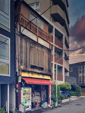 A small, weathered shop is nestled between modern buildings on an urban street. The shop has a faded sign and an old awning, with a vending machine and various items displayed outside. The surrounding structures are tall and contemporary, creating a stark contrast with the traditional shop. The street is empty, and the overall atmosphere suggests a blend of past and present architectural styles.