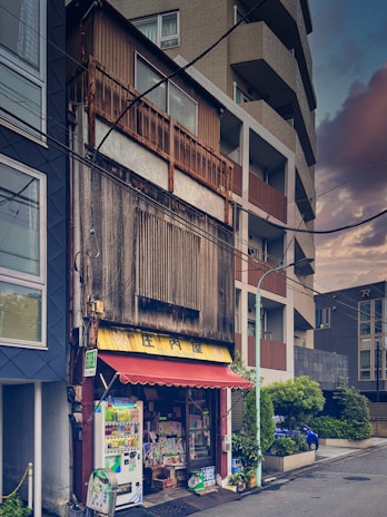 A small, weathered shop is nestled between modern buildings on an urban street. The shop has a faded sign and an old awning, with a vending machine and various items displayed outside. The surrounding structures are tall and contemporary, creating a stark contrast with the traditional shop. The street is empty, and the overall atmosphere suggests a blend of past and present architectural styles.