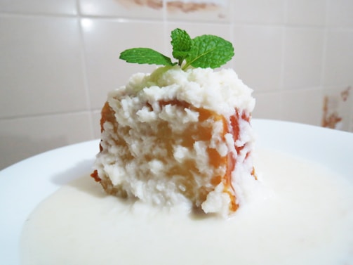 A slice of white, creamy dessert with a textured surface, topped with a fresh mint leaf. The dessert appears to be served on a white plate, with a sauce or cream surrounding its base. The background features neutral-colored tiles.