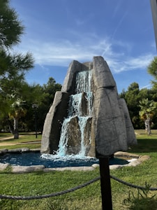 A landscaped garden features an artificial rock waterfall with flowing water cascading down into a small pond. Surrounding the waterfall are well-maintained green lawns, trees, and palm trees under a clear blue sky.
