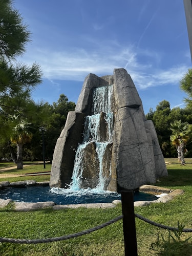 A landscaped garden features an artificial rock waterfall with flowing water cascading down into a small pond. Surrounding the waterfall are well-maintained green lawns, trees, and palm trees under a clear blue sky.