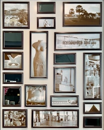A collection of 18 framed pictures arranged on a wall, featuring a variety of subjects including sculptures, bottles, a tea set, and architectural scenes. The photos are black and white, with a mix of both large and small frames. One central image is of a torso sculpture, while other subjects include a teapot, a large pot, and various indoor and outdoor scenes.