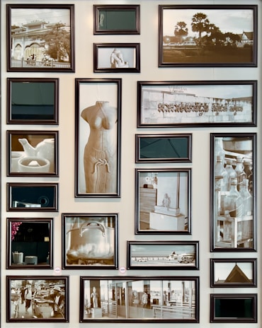 A collection of 18 framed pictures arranged on a wall, featuring a variety of subjects including sculptures, bottles, a tea set, and architectural scenes. The photos are black and white, with a mix of both large and small frames. One central image is of a torso sculpture, while other subjects include a teapot, a large pot, and various indoor and outdoor scenes.