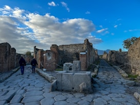 The image depicts an ancient stone-paved street flanked by weathered brick ruins and fragmented structures, with a partly cloudy sky overhead. Two individuals walk on the left side of the street, surrounded by the historical remnants of an archaeological site.