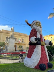 A large, festive Santa Claus figure is prominently displayed in front of a historic building labeled 'Conservatorio di S.M. Delle Grazie.' The building is characterized by its classical architecture and Italian flags. A statue and well-maintained garden with red flowers are also present. The sky is clear, and a few palm leaves are visible on the side.