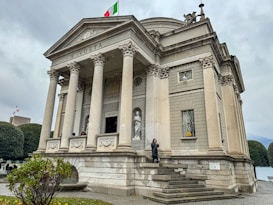 A neoclassical building with a series of tall columns at the entrance, topped with a triangular pediment. There is an Italian flag on top of the building. A small group of people and a statue are visible in front of the entrance. The sky is overcast, and there are neatly trimmed bushes and a small garden in the foreground.