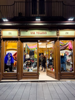 A clothing store display with wooden frames and glass windows showcasing various garments and shoes. Inside, two people are browsing the merchandise. The sign above the entrance reads 'VIA TOLEDO' and the street number 291 appears on both sides of the entrance.