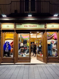 A clothing store display with wooden frames and glass windows showcasing various garments and shoes. Inside, two people are browsing the merchandise. The sign above the entrance reads 'VIA TOLEDO' and the street number 291 appears on both sides of the entrance.