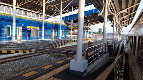 A modern train station with multiple tracks and platforms. The station features a bright blue building with clear signage, surrounded by white pillars and an arched roof. The infrastructure includes railings, stairs, and tactile paving for accessibility. The tracks are clear, and there's a visible train on the right, indicating that the station is operational.