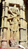 A stone carving of a historical figure is prominently featured, showcasing elaborate details in the sculpture, with intricate facial expressions and clothing design. The figure is situated between two stone pillars and is depicted holding objects in its hands. At the bottom of the sculpture, a white bird stands near the base, adding a contrast to the overall earthy tones of the stone.