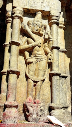 A stone carving of a historical figure is prominently featured, showcasing elaborate details in the sculpture, with intricate facial expressions and clothing design. The figure is situated between two stone pillars and is depicted holding objects in its hands. At the bottom of the sculpture, a white bird stands near the base, adding a contrast to the overall earthy tones of the stone.