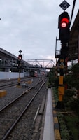 A railway track with a red traffic signal indicating stop. The track forks into two directions. There are multiple overhead pedestrian bridges in the background. The environment appears to be urban with some greenery on one side.