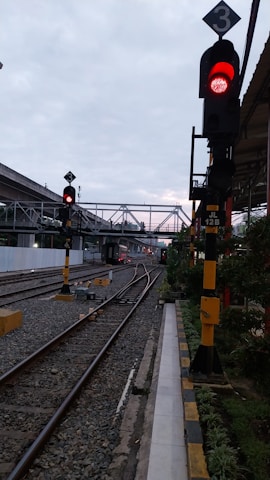 A railway track with a red traffic signal indicating stop. The track forks into two directions. There are multiple overhead pedestrian bridges in the background. The environment appears to be urban with some greenery on one side.